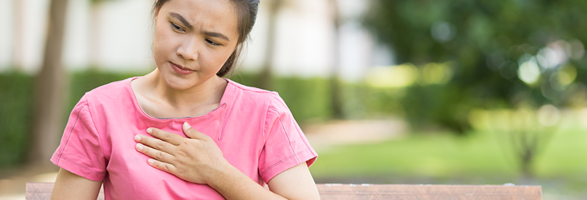 A young woman sits outside and holds her chest as she experiences heartburn.