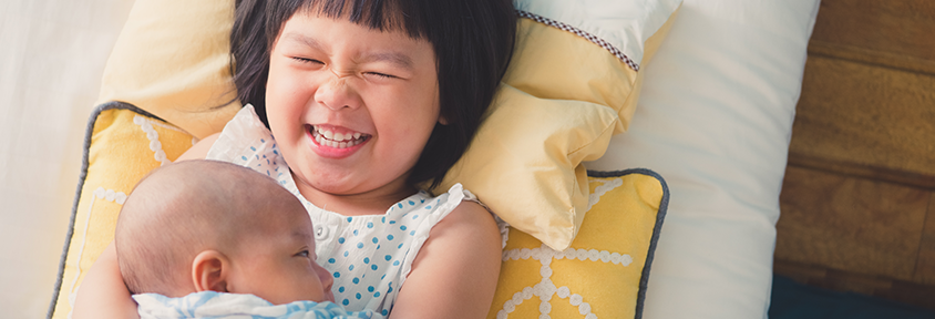 A little girl happily holds her newborn sibling, showing a healthy sibling relationship.