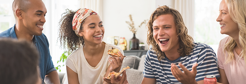 A group of vaccinated friends sits together eating pizza and laughing. 