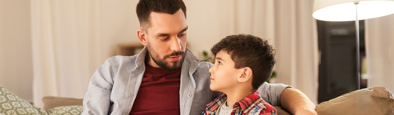 A father seated on a sofa with his child, talking about the importance of mental health.