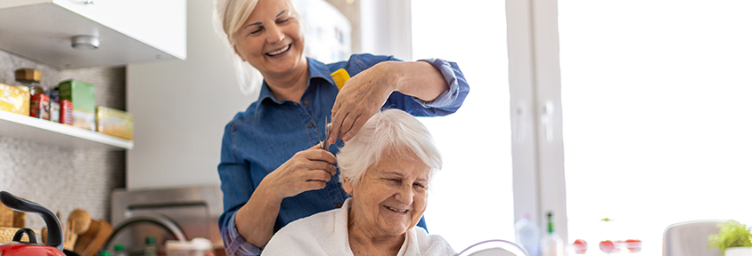 Daughter cuts her senior mother’s hair in the kitchen while she acts as her family caregiver.