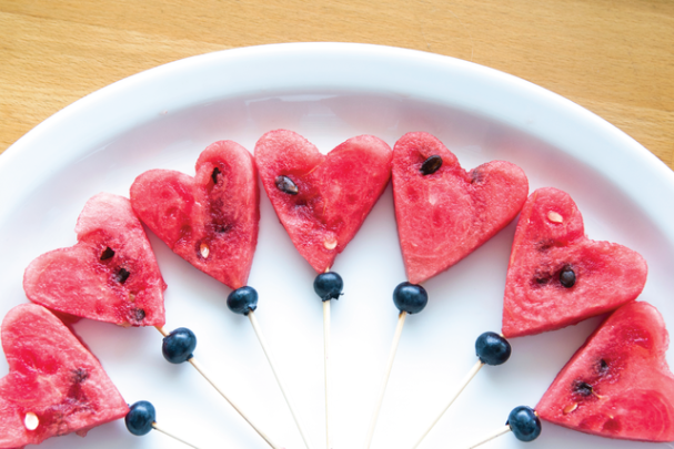 Pieces of heart-shaped watermelon on a spear with a blueberry accent underneath as part of a healthy school lunch.