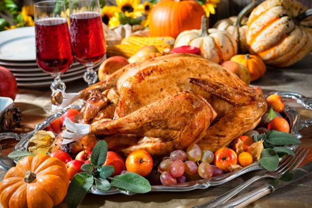 A roasted turkey sits on a platter surrounded by vegetables and glasses of a red beverage