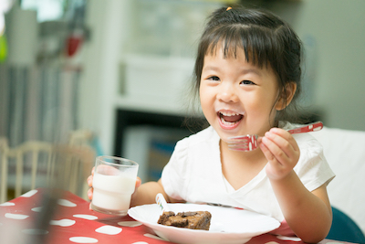 A young girl happily eats a brownie with a glass of milk.