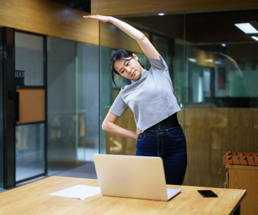A young woman takes a break from working at her desk to stand and stretch.