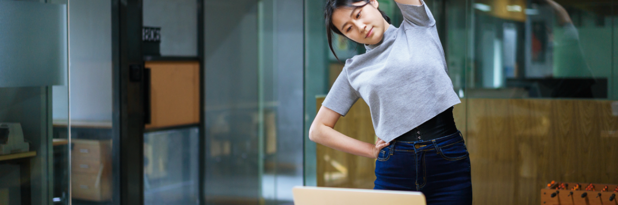 A young woman takes a break from working at her desk to stand and stretch.