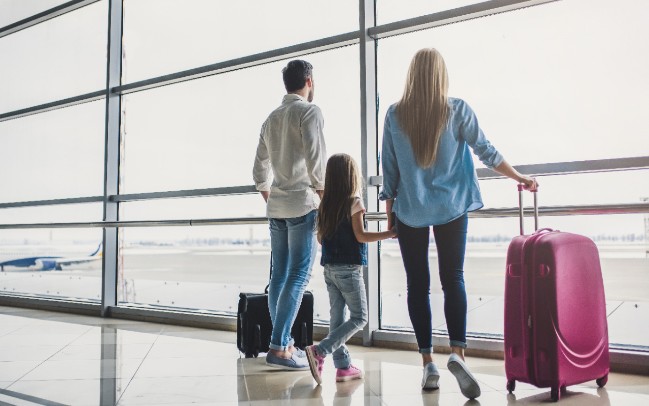 A family stares out an airport window while holding their luggage 