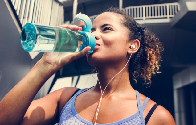 A woman drinks from a water bottle as she gets ready to exercise. 