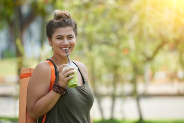 A young woman holds a yoga mat in a park and sips a green smoothie