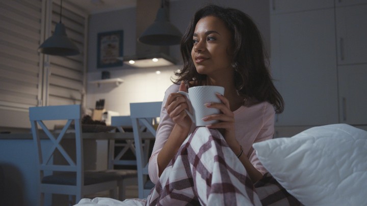 A woman sits in her pajamas in a dark room while drinking out of a mug