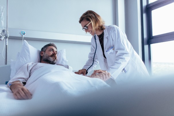 A man lays in a hospital bed while a doctor uses a stethoscope to check his heartbeat