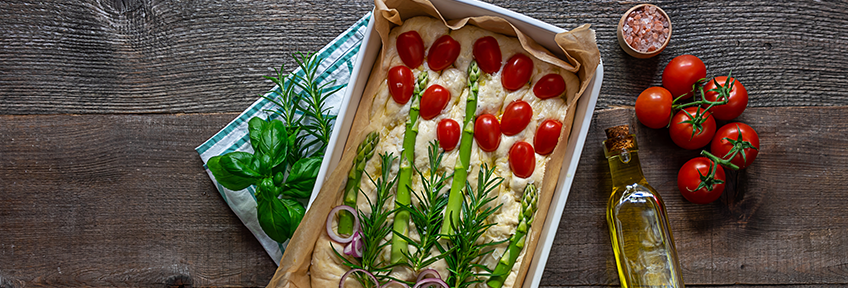 A pan of unbaked focaccia sits on a table with tomatoes and asparagus arranged to look like flowers.