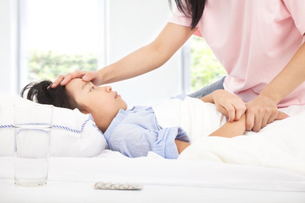 Sick child lies in bed while their mother checks their head for signs of a fever